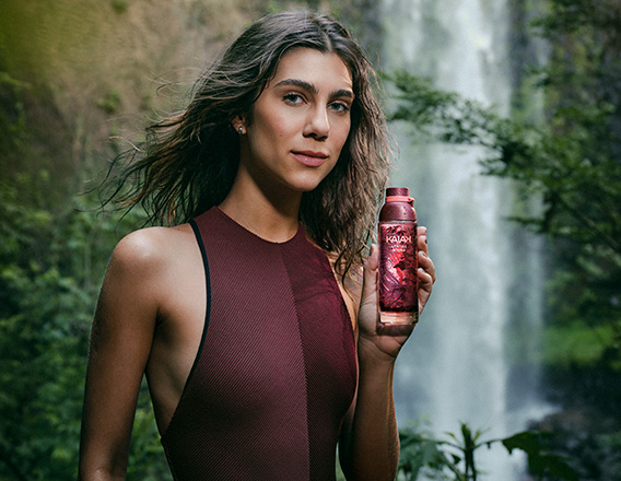 Modelo morena de cabelos lisos e soltos usa maiô vinho e segura o novo Kaiak Aventura Intensa Feminino nas mãos. Ao fundo, destaca-se a imagem de uma cachoeira em meio à floresta.