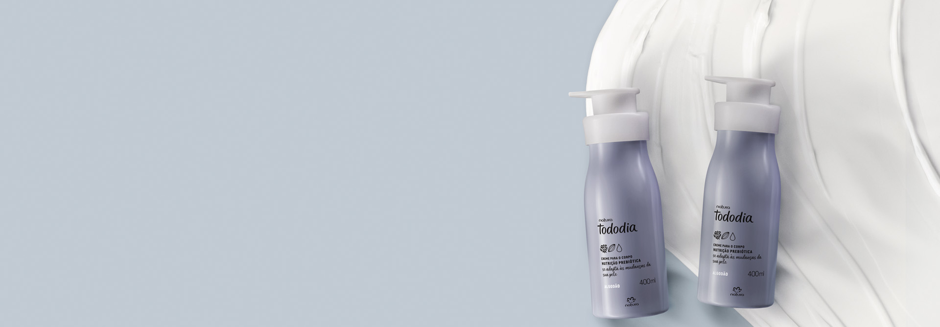 2 frascos pump cinzas do Creme Desodorante Nutritivo Corporal Tododia Algodão deitados sobre um pouco de produto com textura cremosa branca. Fundo azul-claro.