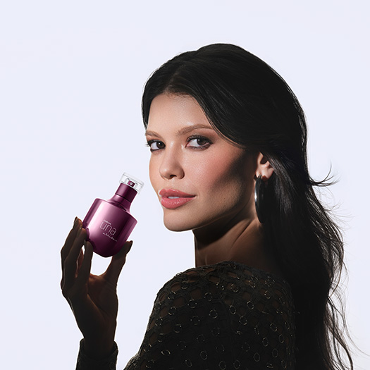 Mulher com maquiagem marcante e cabelos castanhos soltos segura frasco roxo de Natura Una Artisan próximo ao rosto. Ela usa brincos e roupa prateadas, em fundo branco. 