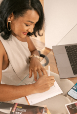 Consultora de Beleza Natura e Avon, de pele morena e cabelos escuros e curtos, está sentada à mesa escrevendo. À sua frente, revistas Natura e Avon, app Natura no celular e site Natura no notebook.