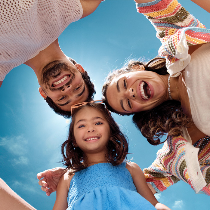 Homem branco de cabelos castanhos veste branco, mulher branca de cabelos castanhos semipresos usa tricô multicolorido com top branco e menina de cabelos castanhos está de vestido azul. O fundo é um céu azul.