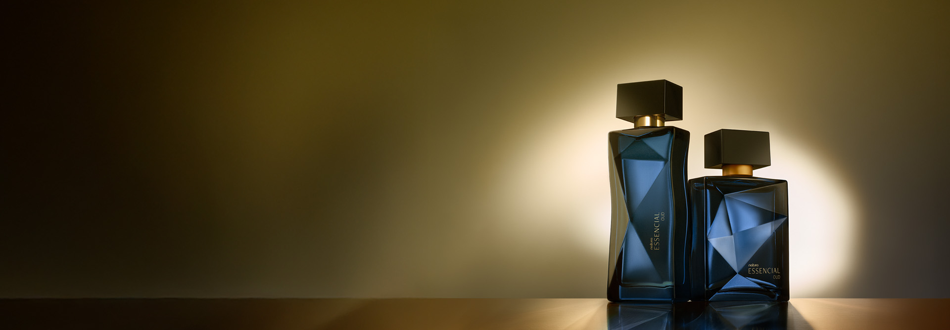 Essencial Oud Feminino e Masculino, com frascos azul-marinho e tampas pretas, aparecem iluminados no canto direito da tela sobre superfície e fundo em tom de marrom.