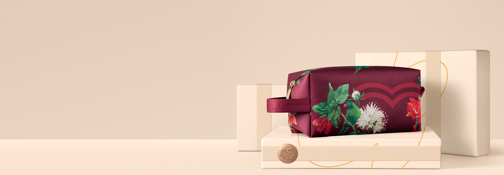 Nécessaire vinho com estampa floral e alça sobre caixas presenteáveis creme com selo dourado. Fundo creme.