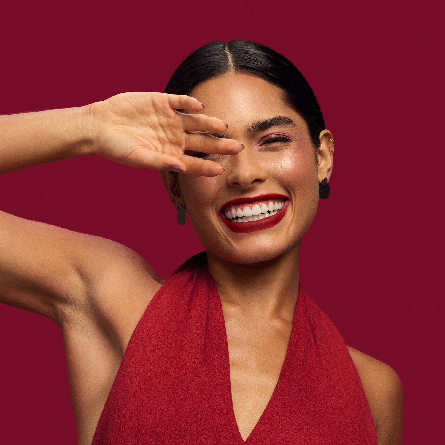 Mulher jovem de pele bronzeada e cabelo preto preso sorri enquanto leva a mão ao rosto. Ela usa um vestido vermelho e batom vermelho vibrante na tom cereja. Fundo vermelho. 