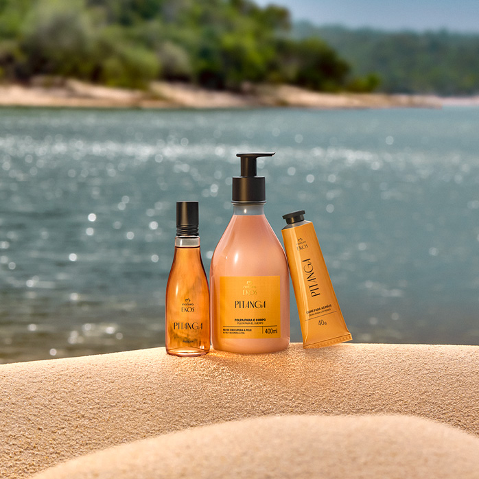 Três produtos Natura Ekos Pitanga em embalagens amarelas e alaranjadas estão sobre areia clara, com rio e vegetação ao fundo, transmitindo sensação de frescor e natureza.
