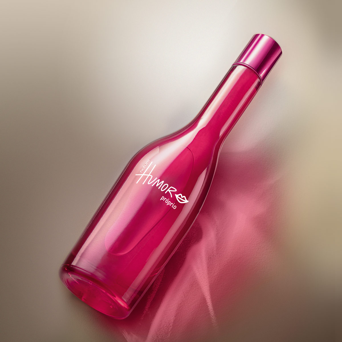Foto do produto deitado, em um fundo marrom metalizado. O Humor Próprio conta com uma embalagem em vidro rosa, arredonda, similar a uma garrafa com um bocal fino e longo. Sua tampa é, também, rosa metalizada. Na embalagem está estampado o nome da colônia Humor Próprio em branco com um desenho em linhas brancas de uma boca.