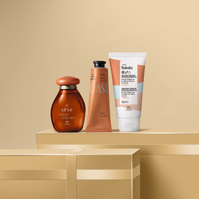 Trio de produtos Natura (Óleo Sève, Creme Ekos e Hidratante Tododia) posicionados sobre caixas de presente douradas em um cenário minimalista.