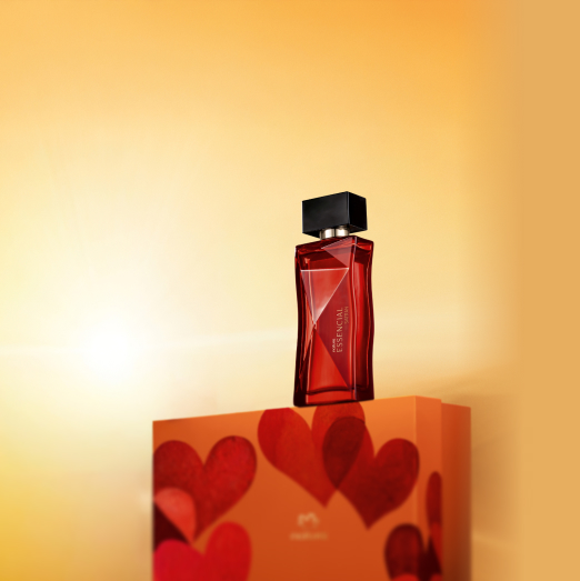 Frasco de vidro vermelho com tampa preta do Deo Parfum Essencial Safran Feminino em destaque sobre caixa de presente laranja com corações vermelhos, em fundo degradê amarelo.