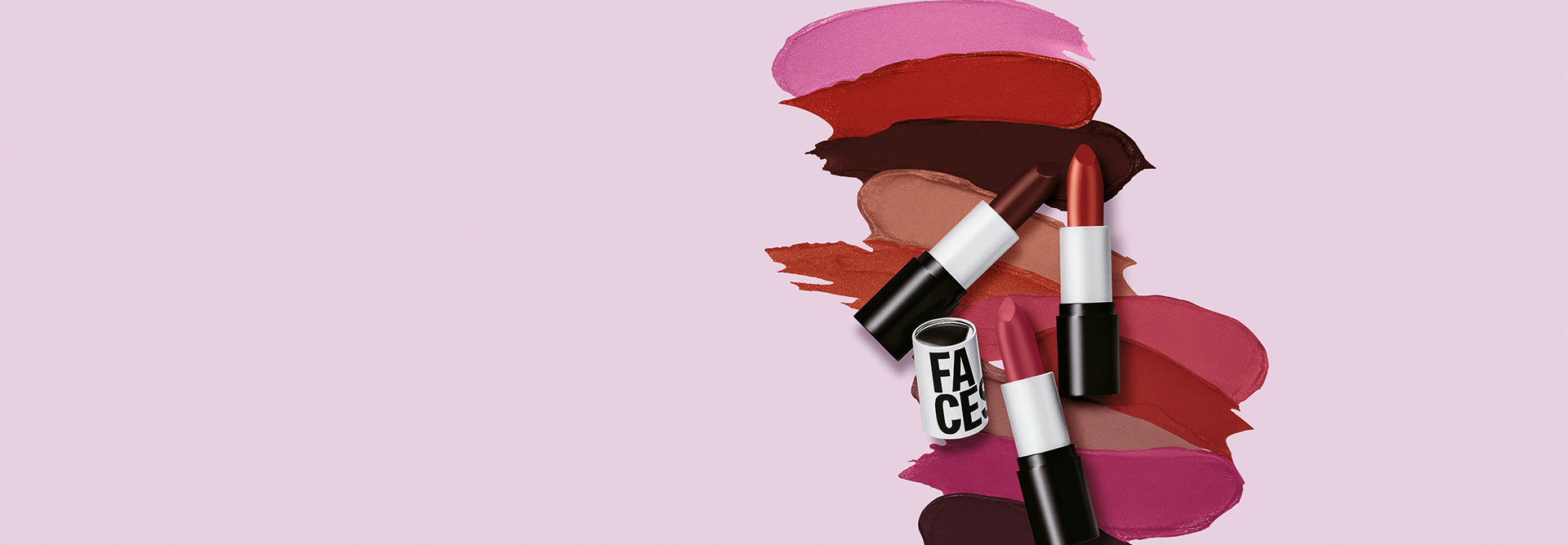 Três batons da linha Natura Faces abertos, exibindo tons de vinho, vermelho e rosa, sobrepostos a diversas manchas de cores de batom que variam entre nudes, rosas e marrons. O fundo é um degradê suave de rosa para cinza.