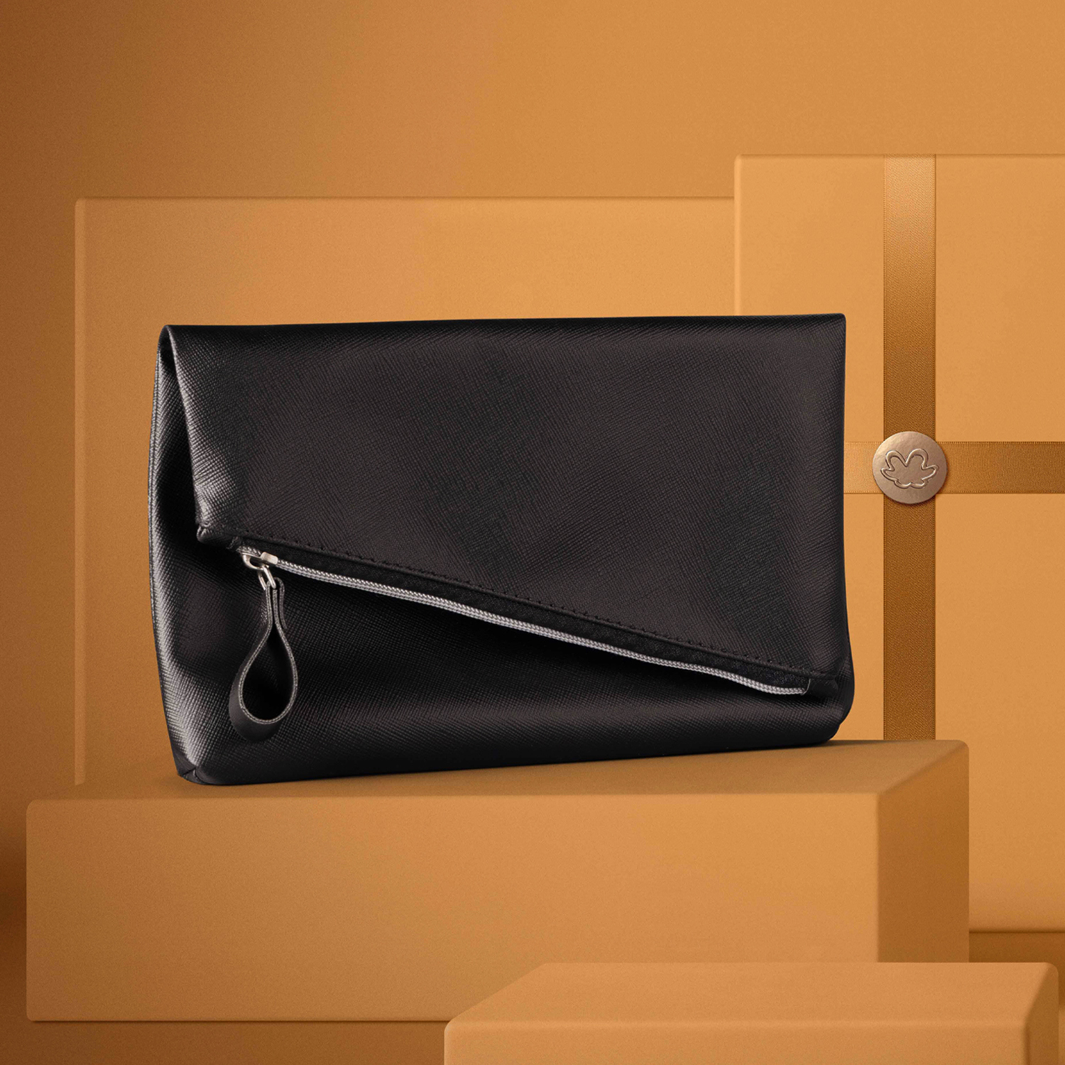Bolsa de mão preta estilo envelope em tecido de couro sintético, com zíper e detalhes em prateado repousa sobre caixas de presente. Caixas e fundo dourados. 