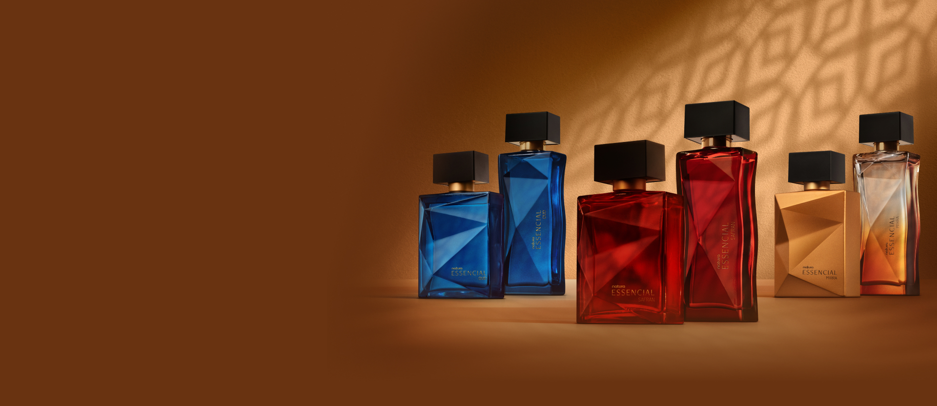 6 frascos de fragrâncias sendo 2 azuis de Essencial Oud, 2 vermelhos de Essencial Safran e 2 alaranjados de Essencial Mirra, todos com tampas pretas. Estão em fundo marrom com sombra geométrica. 