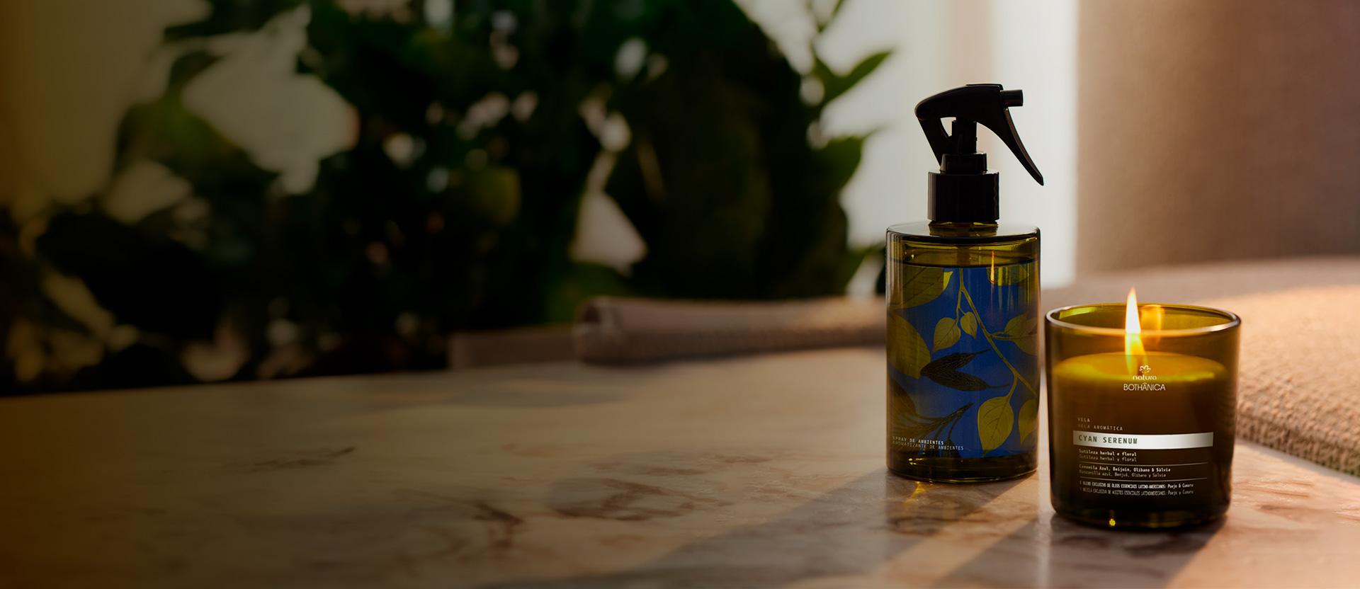 Produtos Natura Bothânica, um spray e uma vela acesa, sobre superfície de mármore com folhagem verde desfocada ao fundo.