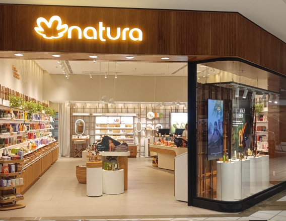 Imagem mostra uma loja física da Natura. Na entrada, logo da Natura iluminado sobre fachada de madeira e vitrine decorada. Dentro da loja, produtos organizados por categorias.