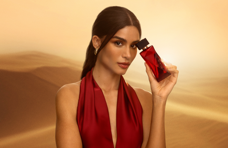 Foto em cenário de dunas ao pôr do sol. Mulher segura o frasco vermelho do perfume Essencial próximo ao rosto.