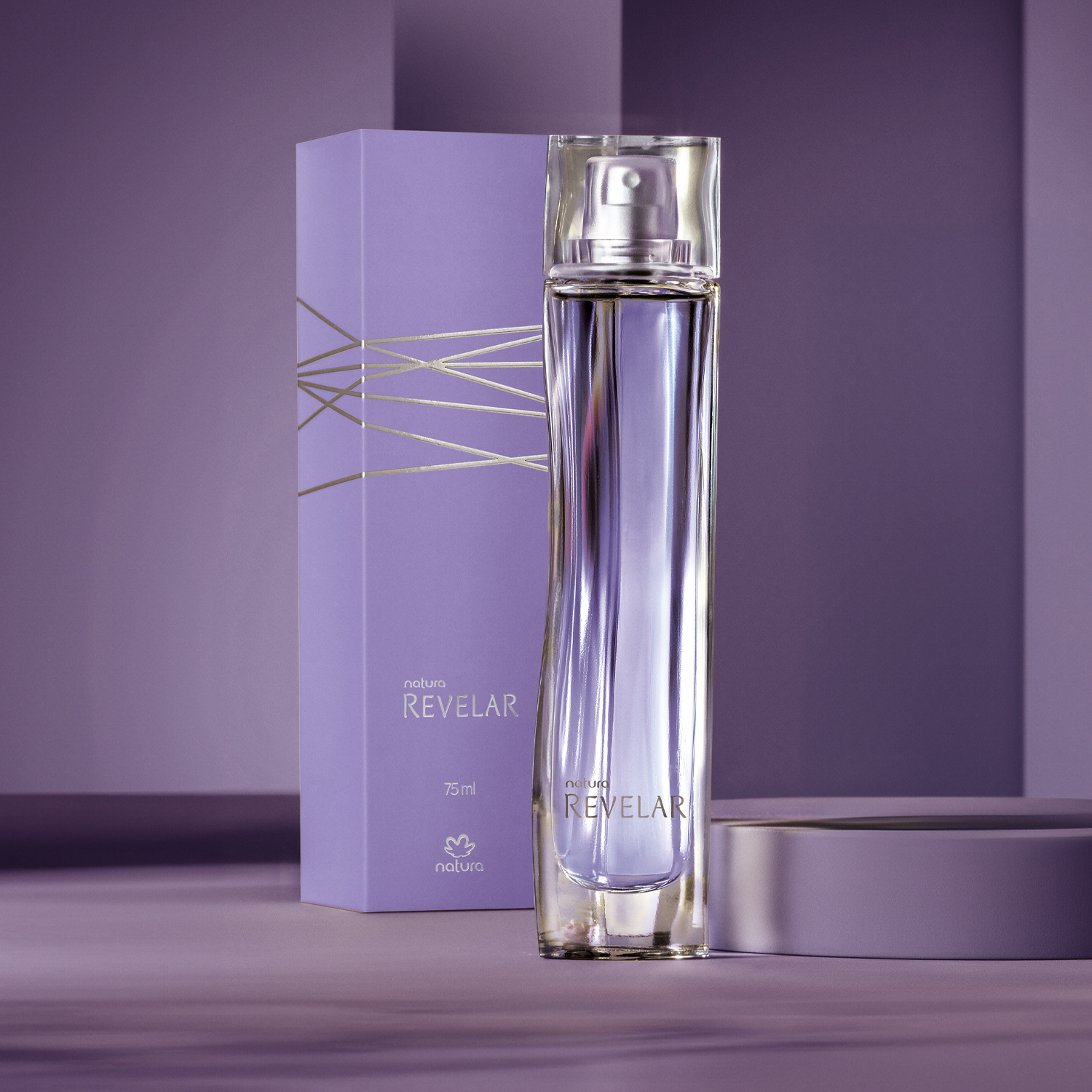O Desodorante Colônia Revelar aparece ao lado da sua caixa roxa com detalhes prateados. O frasco é de vidro transparente, com leve ondulação e tem líquido lilás dentro. Fundo em tons de roxo.