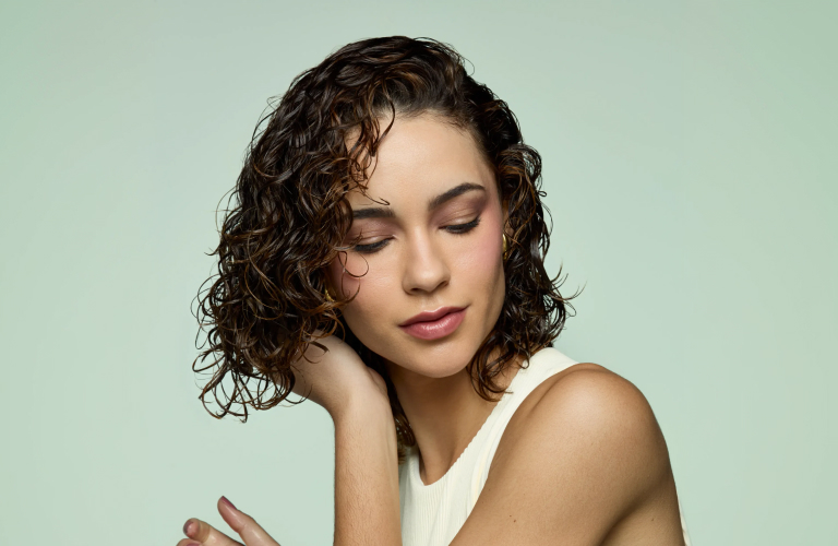 Foto de uma mulher com cabelo cacheado curto, olhos fechados e mão próxima ao rosto. Ao fundo, cor verde claro. Ela veste blusa branca.