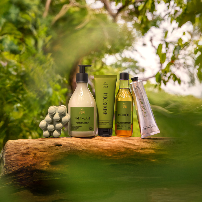 Kit Natura Ekos Andiroba com cinco produtos em embalagens nas cores verdes e bege sobre tronco de madeira. Ao fundo, folhas e árvores desfocadas remetem à natureza exuberante.