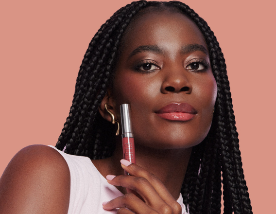 Foto de mulher negra de tranças segurando um batom Natura Una próximo ao rosto, em fundo rosa-claro.