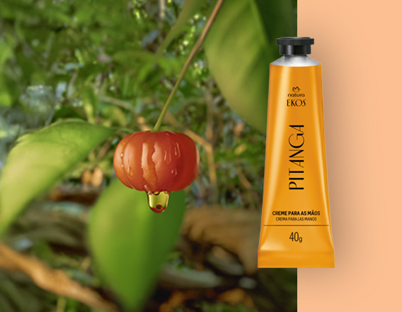 Bisnaga amarela do Creme Hidratante para as Mãos Ekos Pitanga à direita, ao lado de uma pitanga vermelha com gota de extrato, sobre fundo de folhas verdes e laranja suave.