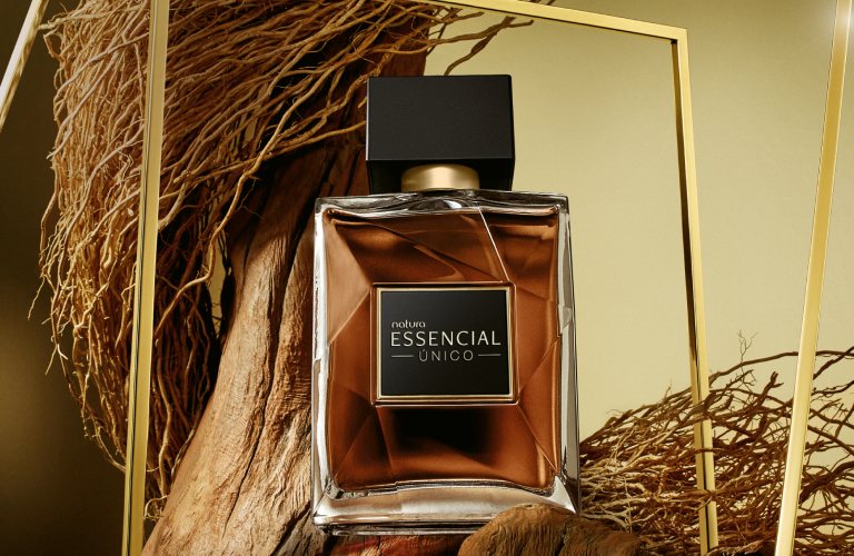  Perfume doce Essencial Único Masculino posicionado em meio a um galho de árvore e cipó. O fundo da imagem marrom e dourado