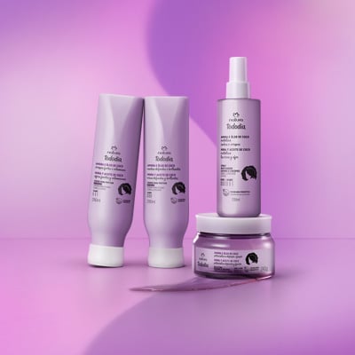 4 produtos da linha Natura Tododia Amora e Óleo de Coco em embalagens lilás. À esquerda, dois tubos de creme estão levemente inclinados; à direita, um frasco de spray está sobre um pote redondo de creme. Uma amostra da textura em gel transparente está espalhada na superfície roxa à frente dos produtos. 