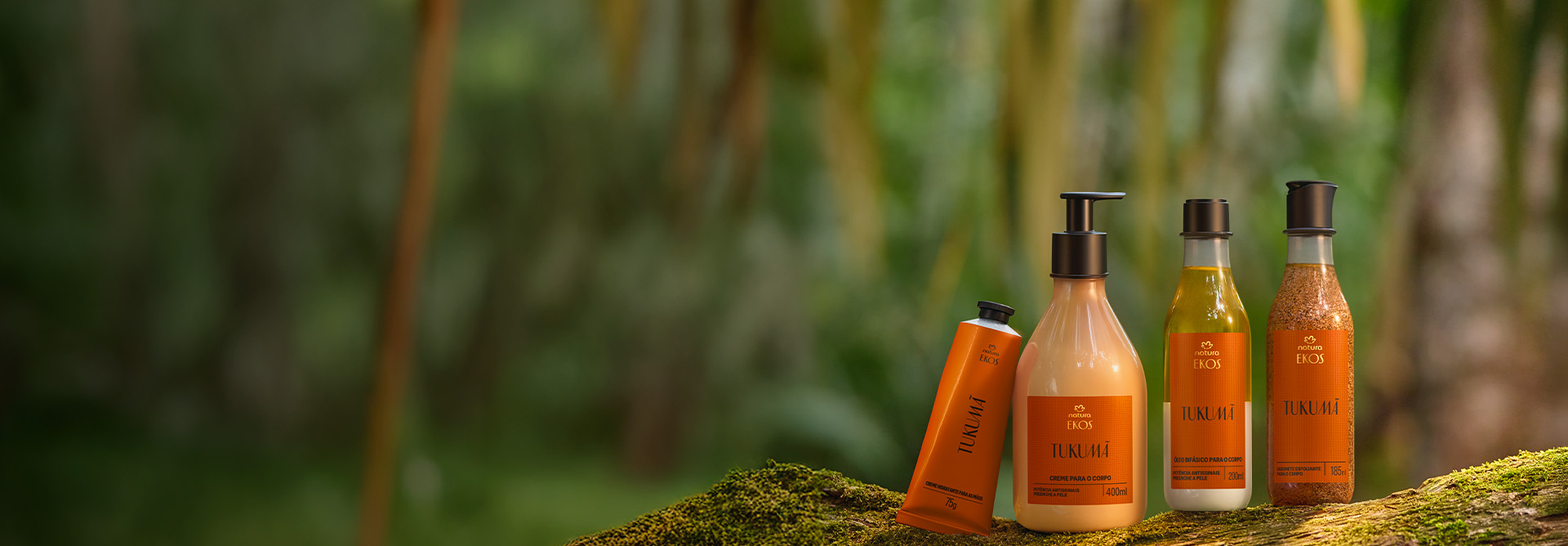 Quatro produtos Natura Ekos Tukumã estão sobre musgo: bisnaga, frasco pump, óleo e esfoliante, todos com rótulo laranja. Ao fundo, floresta desfocada em tons de verde.