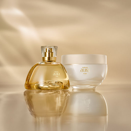 Frasco do Deo Parfum ao lado do Creme Corporal Natura Aura Alba sobre uma superfície dourada, com fundo suavemente iluminado. As embalagens são elegantes com detalhes em dourado.