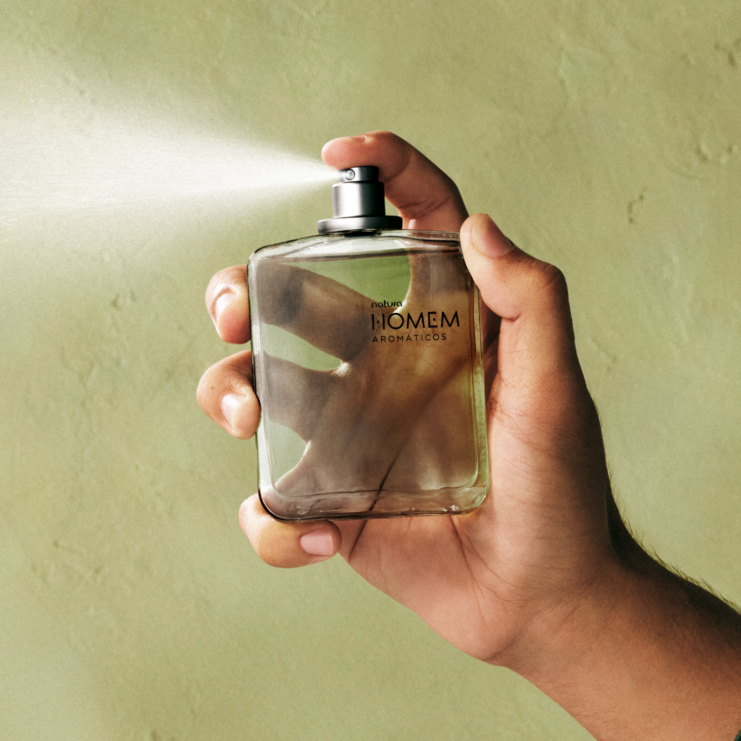 Foto de uma mão masculina que segura o frasco retangular do Desodorante Colônia Natura Homem Aromáticos, borrifando a fragrância. Fundo da imagem é verde.