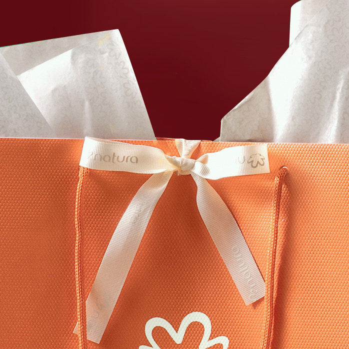 Sacola de presente da Natura com laço e papel de seda saindo da sacola. A sacola está sobre fundo vermelho.