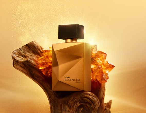 Deo Parfum Essencial Mirra Masculino em frasco facetado dourado com tampa preta. Está em fundo areia sobre um tronco de madeira escura e pedras âmbar ao redor.