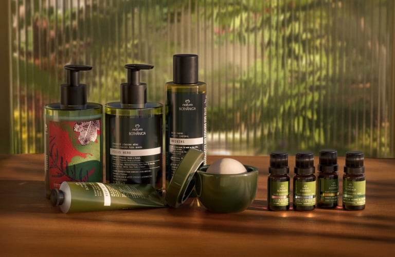 9 produtos de Bothânica, incluindo pump de sabonete, bisnaga de creme, óleo corporal, difusor cerâmico e 4 óleos essenciais.  Embalagens verdes de vidro sobre sobre uma mesa de madeira, com vidro canelado ao fundo.