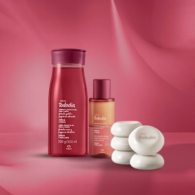 Sabonete Líquido para o Corpo, Óleo Bifásico e Sabonete em Barra Tododia Cereja e Avelã sobre fundo vermelho. O frasco do sabonete líquido é vermelho, do óleo bifásico é transparente e os sabonetes são brancos. 