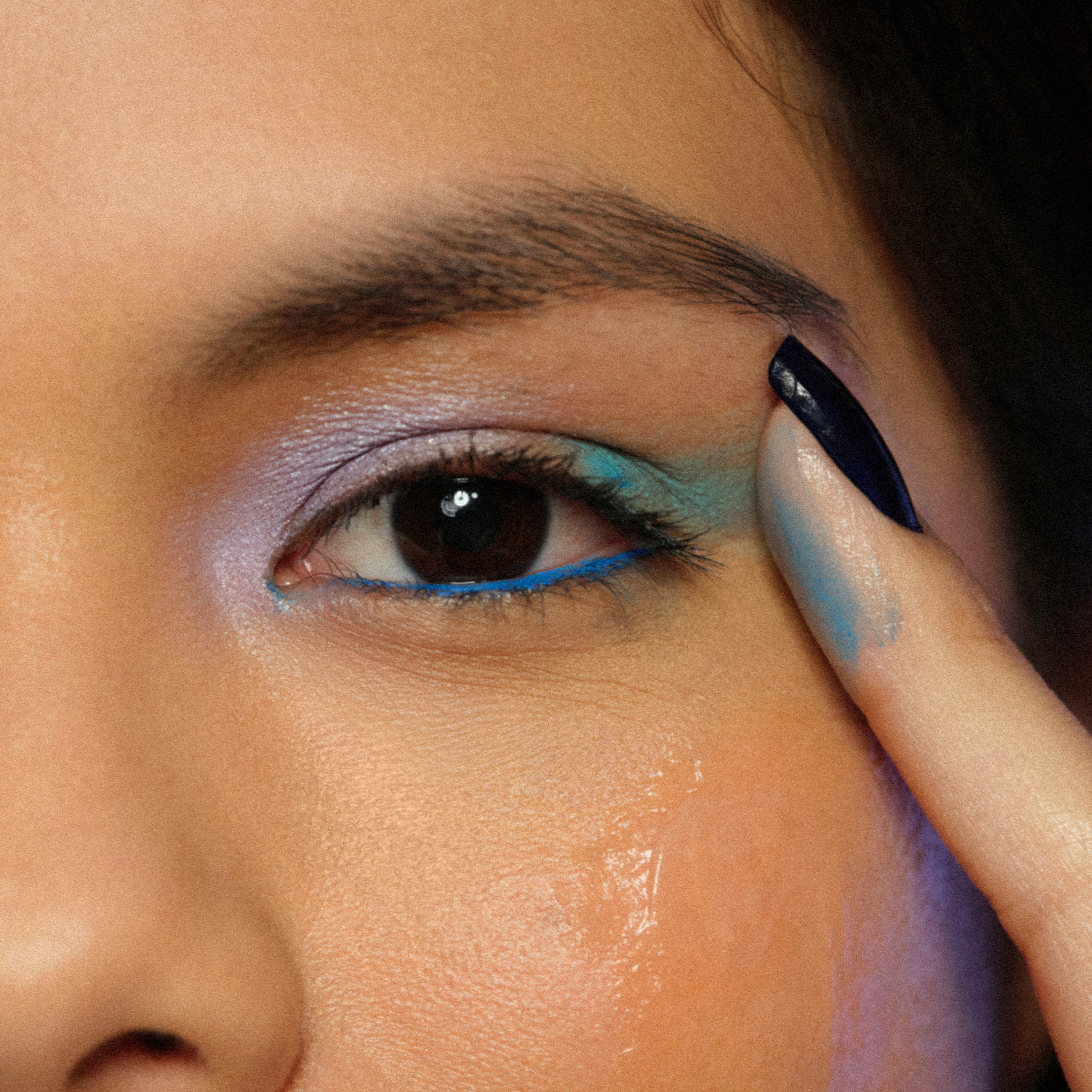 Close nos olhos de uma mulher jovem de cabelo castanho. Ela aplica o Bálsamo Demaquilante Faces sobre o delineado colorido na cor azul, que facilmente é removido pela ação do produto. 
