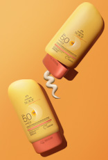 Duas embalagens de protetor solar Natura Solar FPS 50 infantil. Ambas são amarelas com detalhes em laranja avermelhado. Uma das embalagens está deitada na horizontal, com a tampa aberta e o protetor solar branco cremoso espremido para fora em um formato ondulado. A outra embalagem está posicionada verticalmente ao lado dela. O fundo é de um tom laranja vibrante.