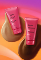 Bisnagas rosa de dois tons de Iluminador Tododia Acerola e Hibisco sobre duas texturas em marrom, claro e escuro, em fundo em tons de amarelo, laranja e rosa.