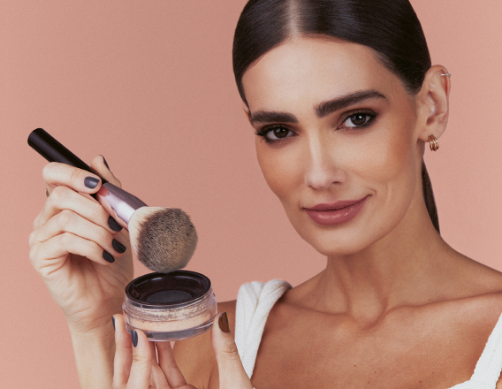 Foto de mulher de cabelos castanhos e presos segurando um produto Natura Una com um pincel, em fundo rosa-claro.