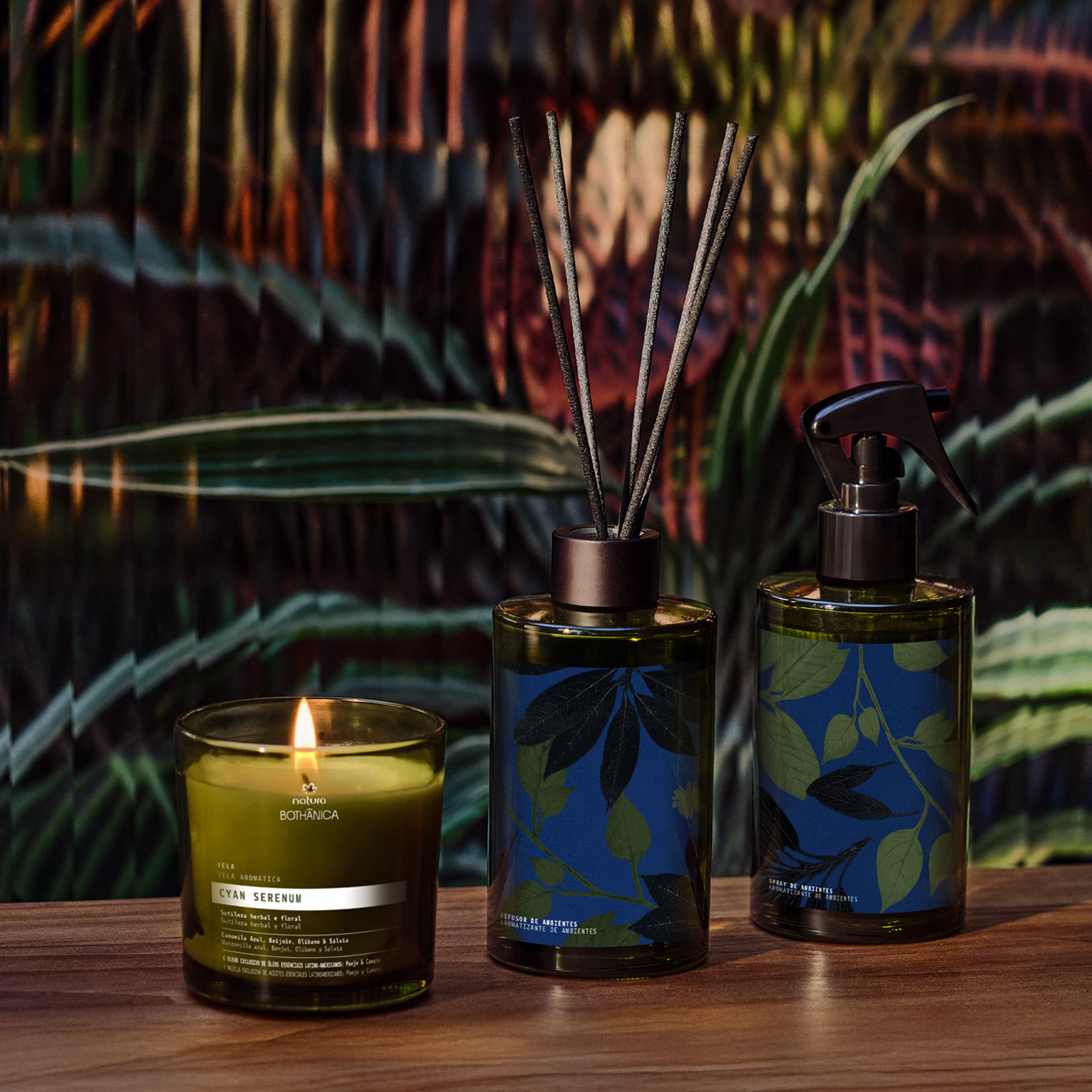 Sobre uma mesa de madeira estão 3 produtos Natura Bothânica Cyan Serenum: uma vela, 1 difusor de ambientes e 1 spray de ambientes. Embalagens em tom verde escuro com detalhes em preto e azul.