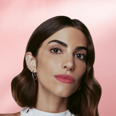 Mulher com cabelos castanhos ondulados na altura dos ombros, usando blusa branca de gola alta. Fundo degradê rosa suave.