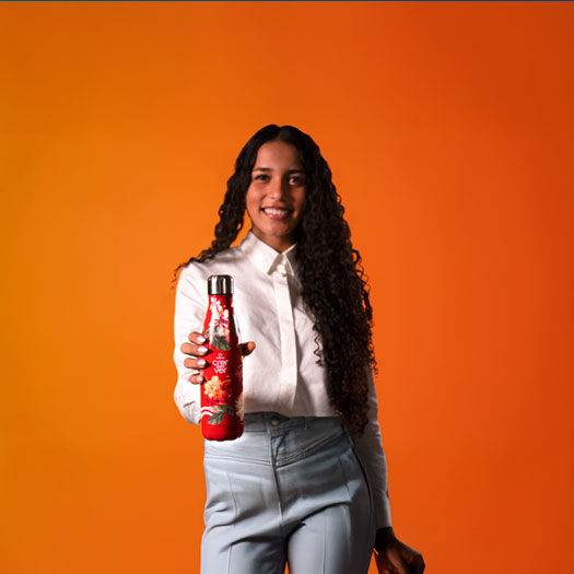 Jovem sorridente com longos cabelos encaracolados, vestindo camisa branca, oferece uma garrafa térmica vermelha com estampa floral vibrante. Fundo laranja liso.