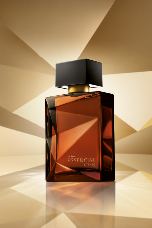 Frasco de Essencial Intenso Masculino, em tons de âmbar e marrom e tampa preta, aparece em destaque em fundo dourado multifacetado, fazendo alusão ao design dos frascos de Essencial.