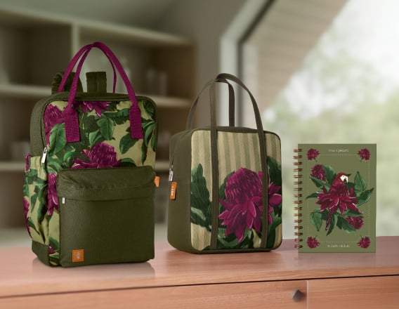 Mochila, frasqueira térmica e caderno Crer Para Ver, todos em tons de verde com estampas de folhagens e flores em tons de roxo e branco, repousam sobre uma bancada de madeira de uma sala com janelas.