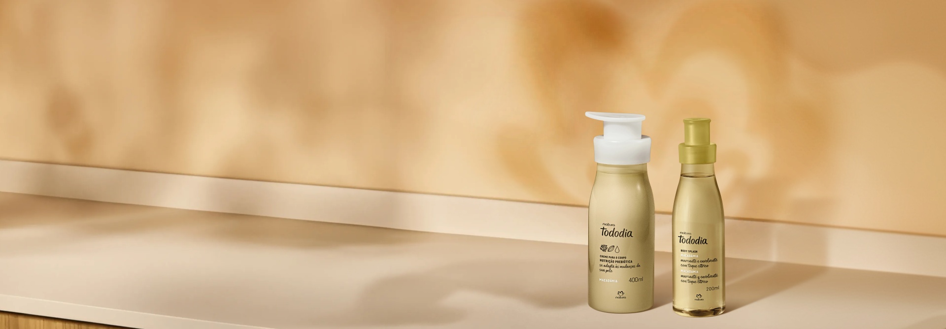 Dois produtos de Tododia Macadâmia sobre um armário branco com portas de madeira, sendo 1 Creme Desodorante Nutritivo para o Corpo e 1 Body Splash. Embalagens em tons de amarelo suave.