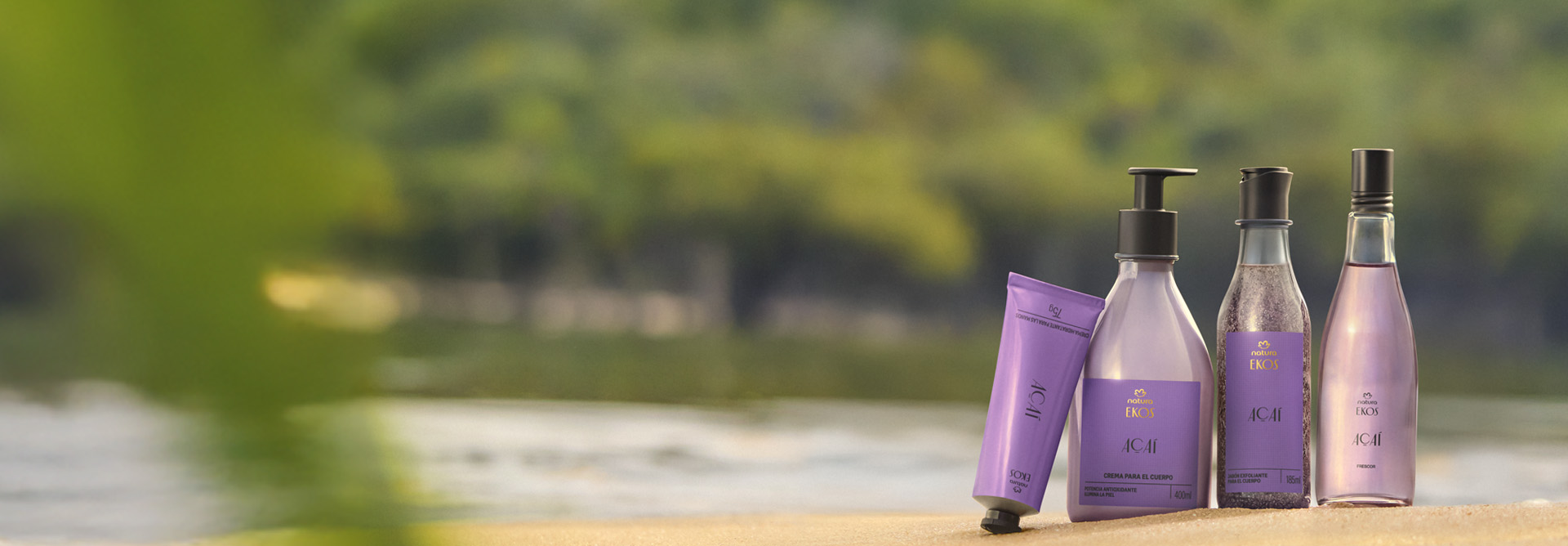 Quatro produtos Natura Ekos Açaí em embalagens lilás estão sobre a areia, com bisnaga, frasco pump, esfoliante e frasco de vidro, em frente a rio e floresta ao fundo desfocado.