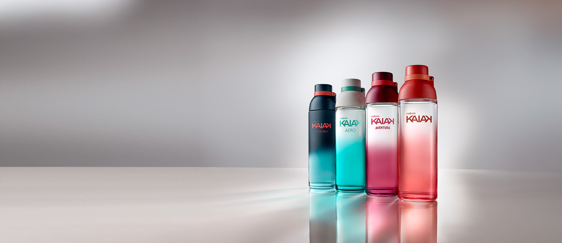 Frascos de Natura Kaiak: Oceano em azul-escuro, Aero em verde-água, Aventura em rosa e Kaiak em vermelho degradê sobre superfície reflexiva com fundo cinza-claro.