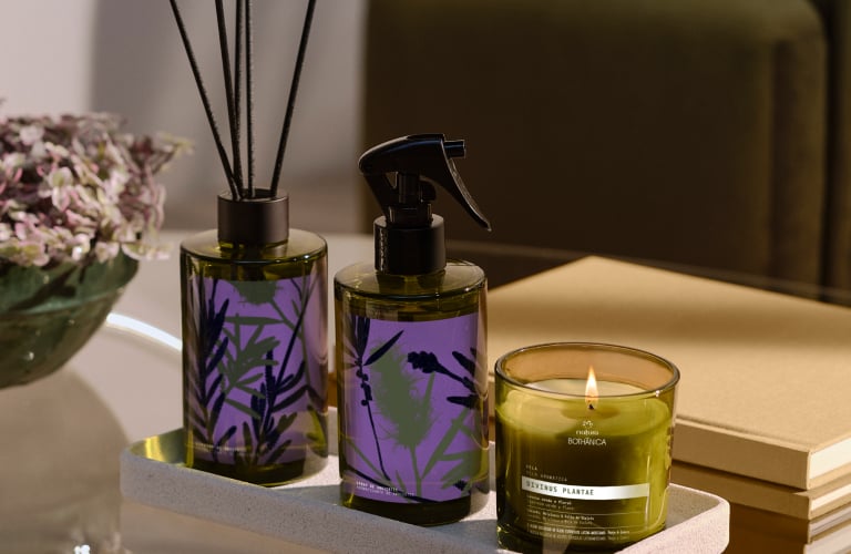 Produtos Bothânica Divinus Plantae: difusor com varetas, spray de ambientes e vela aromática. Embalagens verdes com rótulo roxo em uma bandeja de pedra sobre mesa de vidro, com poltrona verde ao lado.