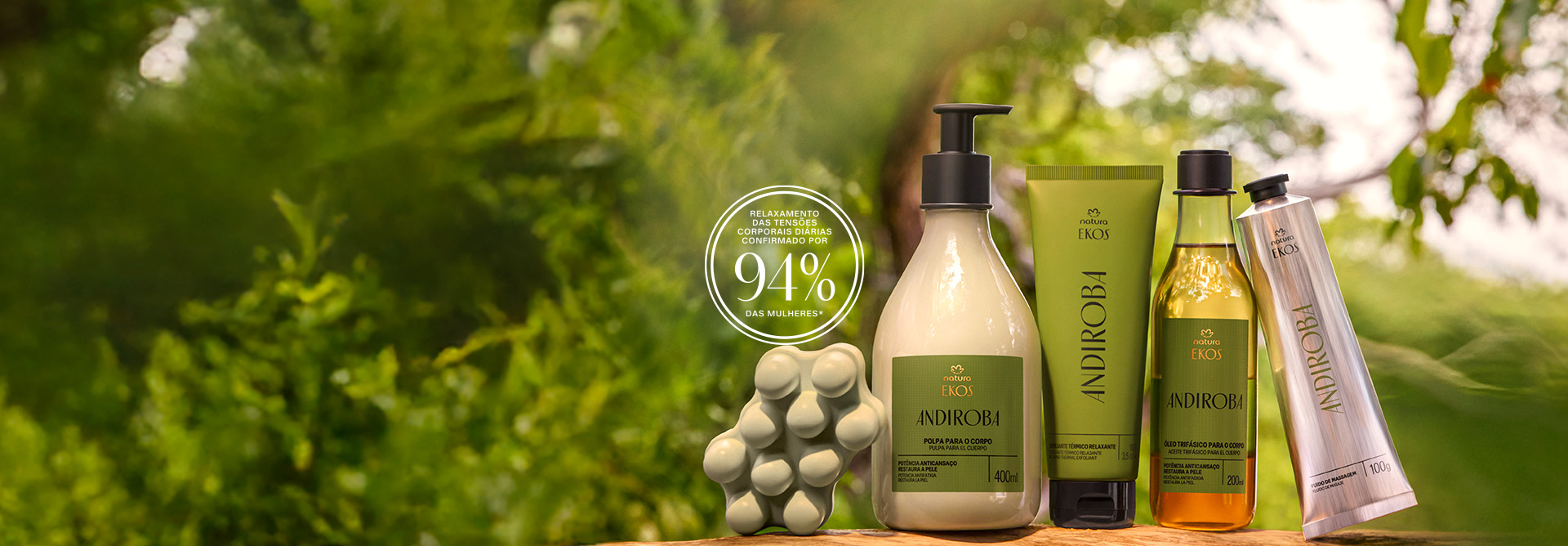 Itens Ekos Andiroba sobre madeira, em mata desfocada: Sabonete em Barra verde, frasco com válvula de Polpa para o Corpo, bisnaga verde de Esfoliante, frasco de Óleo e bisnaga de Fluido de Massagem. Na imagem há um selo com letras brancas e os dizeres: “relaxamento das tensões corporais diárias confirmado por 94% das mulheres*”.