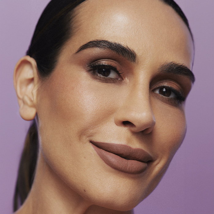 Modelo negra de pele clara, cabelos penteados para trás e pele do rosto uniforme com acabamento natural. A maquiagem com brilho suave, em tons de marrom e nude, realça sua beleza de forma discreta.