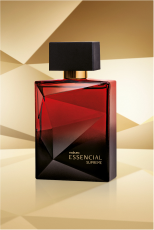 Frasco de Essencial Supreme Masculino, em tom de vermelho e preto e tampa preta, aparece iluminado sobre superfície e fundo dourados.