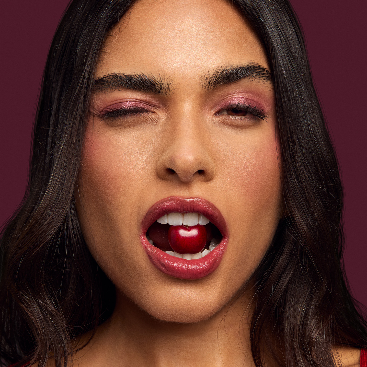 Mulher jovem de cabelo castanho ondulado pisca com um dos olhos e segura entre os dentes uma cereja. Ela usa maquiagem iluminada rosa, gloss nos lábios e blusa vermelha. Fundo roxo. 