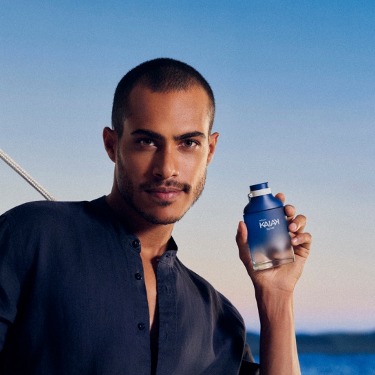 Modelo negro de cabelos curtos e camisa azul-marinho em destaque no centro da tela, ao ar-livre, segurando o Desodorante Colônia Kaiak Sonar Masculino na mão esquerda.